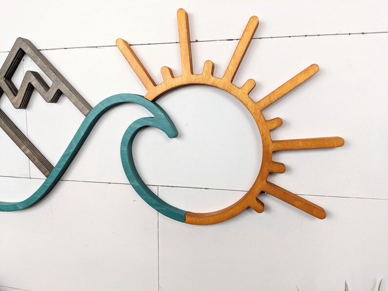 May include: Wooden wall art featuring a mountain range, a teal wave, and a sunburst design. The mountain is gray, the wave is teal, and the sun is a warm wood tone. The art is mounted on a white, horizontal-planked wall.