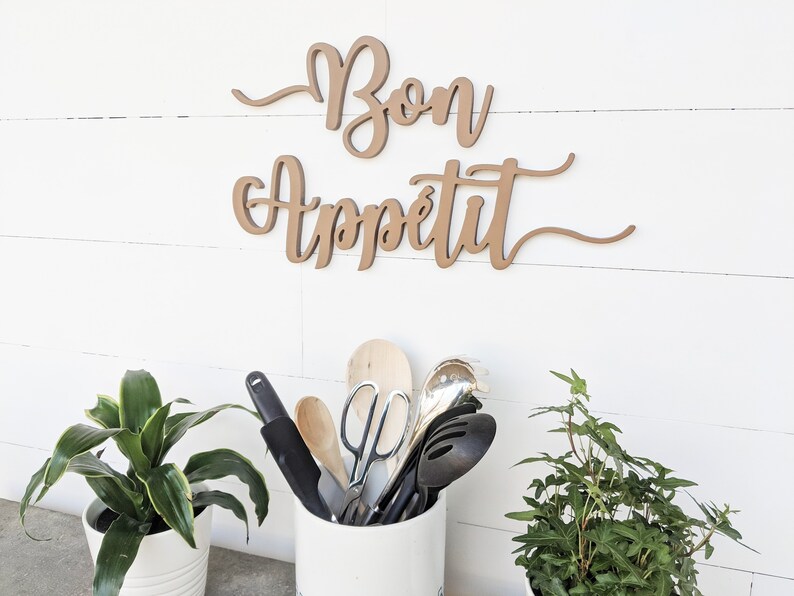May include: A wooden sign with the text 'Bon Appetit' in brown cursive lettering. The sign is mounted on a white wall. In the foreground, a white ceramic utensil holder with a variety of kitchen utensils, including wooden spoons, a spatula, and a pair of kitchen shears. Two potted plants, one with green leaves and the other with dark green leaves, are on either side of the utensil holder.