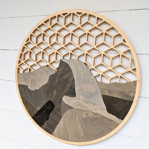 May include: A circular wooden wall art piece featuring a geometric pattern and a mountain scene. The mountain is made of different shades of brown wood, creating a layered effect. The geometric pattern is made of light wood and is cut out of the circle, creating a window-like effect.