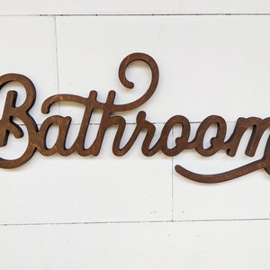 May include: A wooden sign with the word "Bathroom" in a cursive font. The sign is made of dark brown wood and is mounted on a white, wooden plank background. The sign is a decorative item for a bathroom.