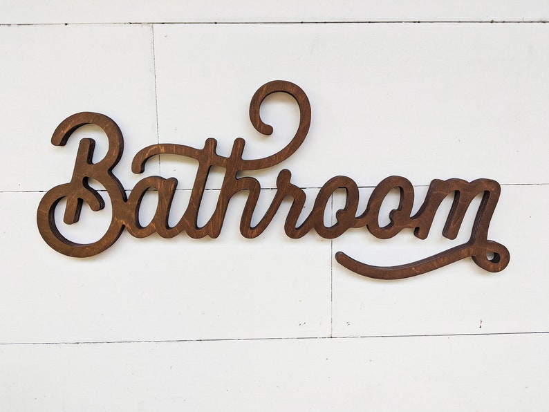 May include: A wooden sign with the word "Bathroom" in a cursive font. The sign is made of dark brown wood and is mounted on a white, wooden plank background. The sign is a decorative item for a bathroom.