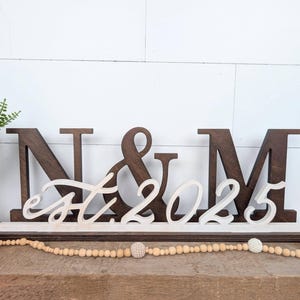 Può includere: Cartello in legno con le lettere "N & M" e la scritta "est. 2025" in corsivo. Le lettere sono marrone scuro e il testo è bianco. Il cartello poggia su una base bianca e marrone, con una ghirlanda decorativa davanti.