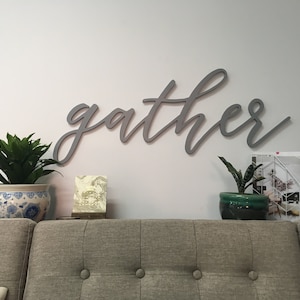 May include: A gray wooden sign with the word "gather" written in cursive script. The sign is hanging on a white wall above a gray couch. There are two potted plants on the table in front of the couch.