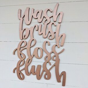 May include: Rose gold colored wooden sign with the words "Wash, Brush, Floss, Flush" written in a cursive font.