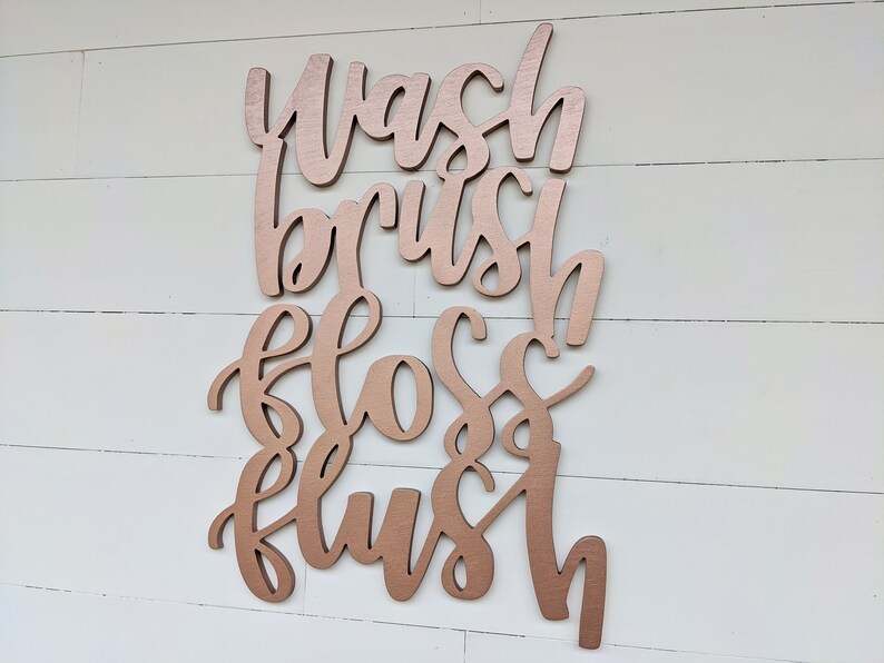 May include: Rose gold colored wooden sign with the words "Wash, Brush, Floss, Flush" written in a cursive font.