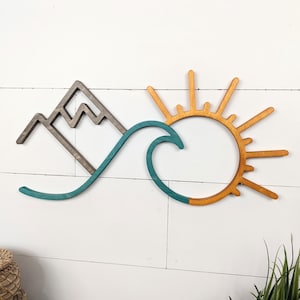 May include: Wooden wall art featuring a mountain, wave, and sun design. The mountain is a dark gray, the wave is teal, and the sun is a warm orange. The art is mounted on a white, horizontal-slat background.