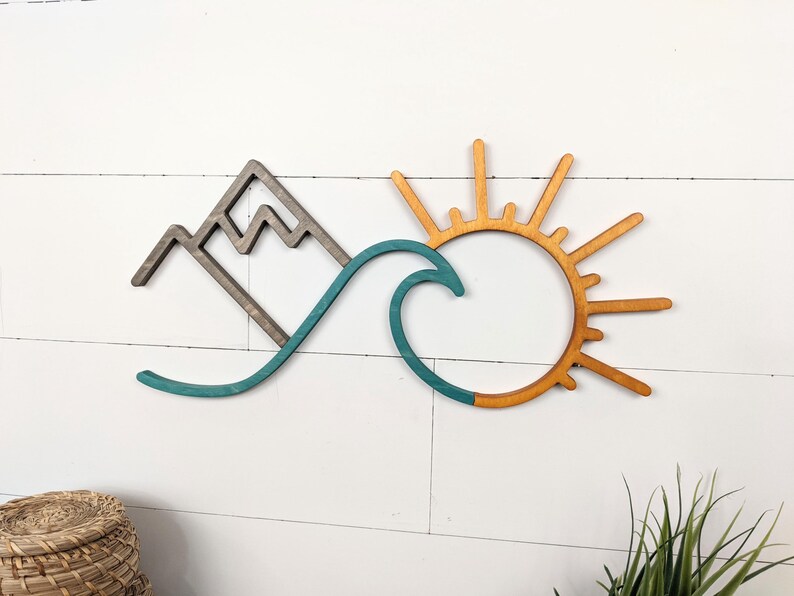 May include: Wooden wall art featuring a mountain, wave, and sun design. The mountain is a dark gray, the wave is teal, and the sun is a warm orange. The art is mounted on a white, horizontal-slat background.