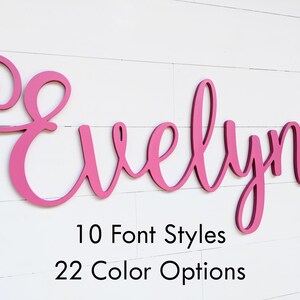 May include: A pink wooden sign spelling "Evelyn" in a cursive font, mounted on a white shiplap wall. The sign is in a vibrant pink color. Below the name are the words "10 Font Styles" and "22 Color Options".
