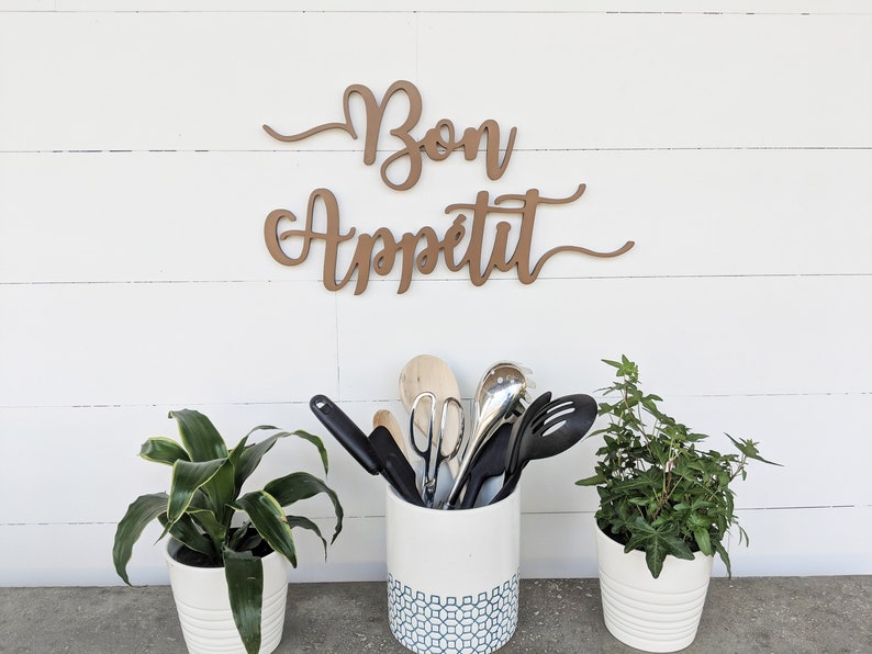 May include: A white ceramic utensil holder with a blue geometric pattern, filled with kitchen utensils, including wooden spoons, spatulas, and tongs. Two potted plants, one with green leaves and the other with small green leaves, are on either side of the holder. A wooden sign with the text "Bon Appetit" is mounted on a white wall behind the holder.