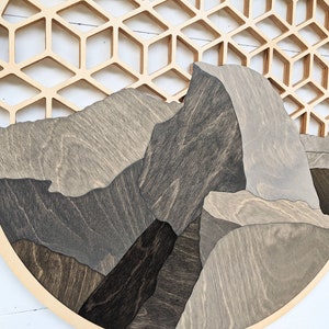 May include: A circular wooden wall art piece featuring a geometric honeycomb pattern in the background and a layered mountain scene in the foreground. The mountains are made of different shades of brown and gray wood.