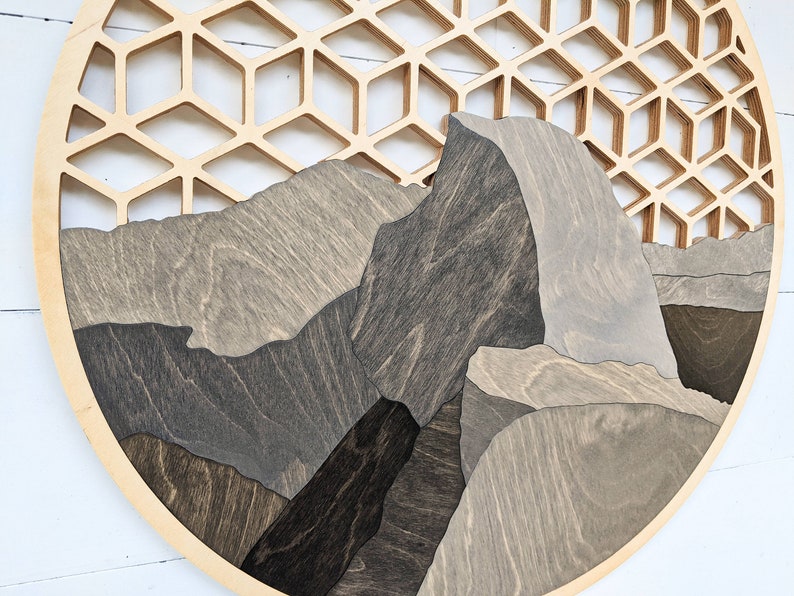 May include: A circular wooden wall art piece featuring a geometric honeycomb pattern in the background and a layered mountain scene in the foreground. The mountains are made of different shades of brown and gray wood.