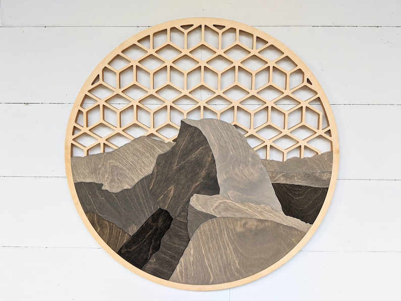 May include: A circular wooden wall art piece with a geometric pattern of hexagons. The center of the piece features a stylized mountain range in shades of brown and gray.