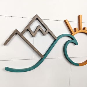 May include: Wooden wall art featuring a mountain range, a teal wave, and a sun with orange and yellow rays. The mountain is brown, and the sun has a circular center. The art is mounted on a white, horizontal-planked wall.