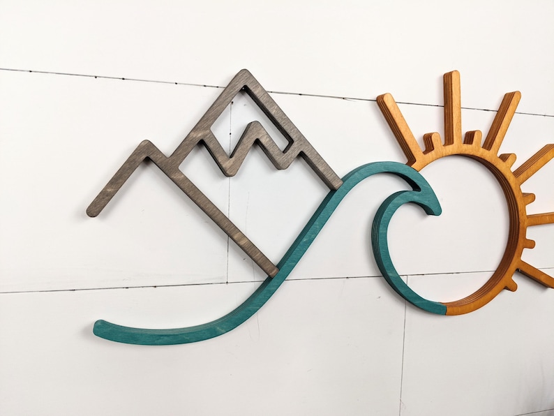 May include: Wooden wall art featuring a mountain range, a teal wave, and a sun with orange and yellow rays. The mountain is brown, and the sun has a circular center. The art is mounted on a white, horizontal-planked wall.