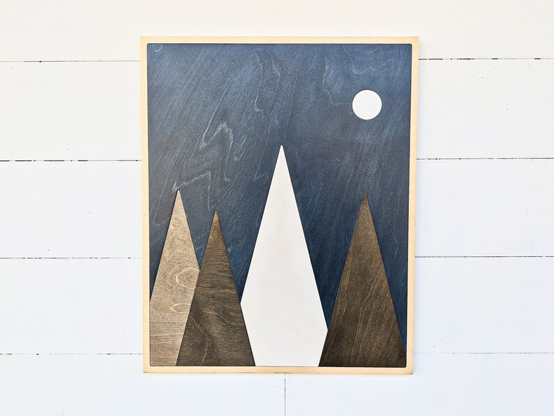 May include: A wooden wall art piece depicting a mountain range with a white moon in a dark blue sky. The mountains are made of different shades of brown wood.