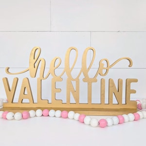 May include: A wooden sign that reads "hello VALENTINE" in gold lettering. The sign is on a white surface with a pink, white, and wooden bead garland in front of it. The background is a white shiplap wall.