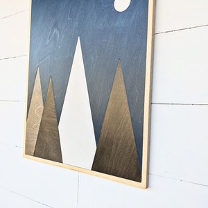 May include: A framed wood wall art piece with a geometric design of three mountains in shades of brown and white against a dark blue background. The mountains are stylized and have a simple, modern look.