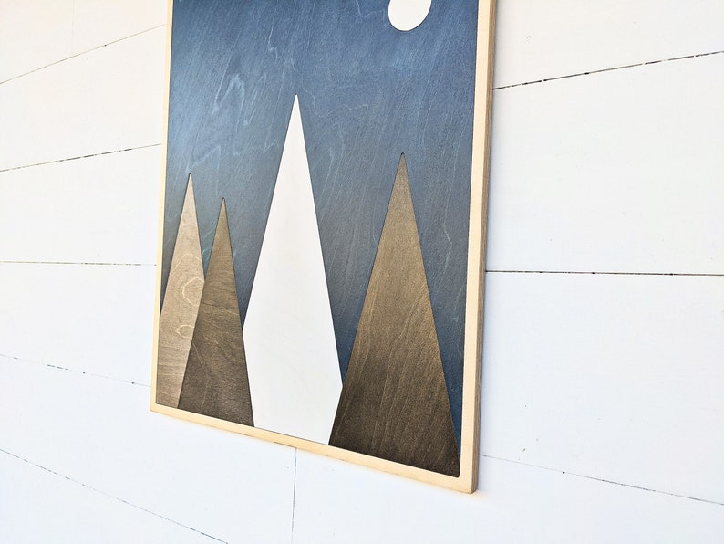 May include: A framed wood wall art piece with a geometric design of three mountains in shades of brown and white against a dark blue background. The mountains are stylized and have a simple, modern look.