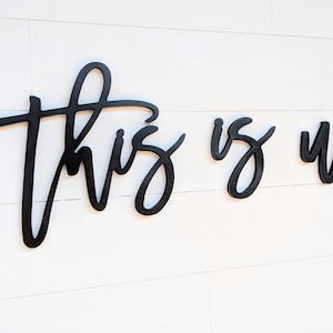 May include: Black wooden wall art spelling out "This is us" in a cursive font. The letters are separate and mounted on a white, horizontal-planked wall. The artwork is a decorative home accent.