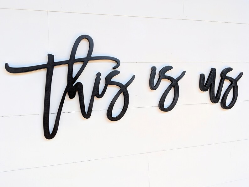May include: Black wooden wall art spelling out "This is us" in a cursive font. The letters are separate and mounted on a white, horizontal-planked wall. The artwork is a decorative home accent.