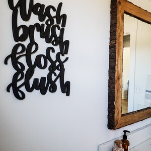 May include: Black wooden sign with the words "Wash, Brush, Floss, Flush" in a stylized font. The sign is hanging on a white wall next to a wooden framed mirror.