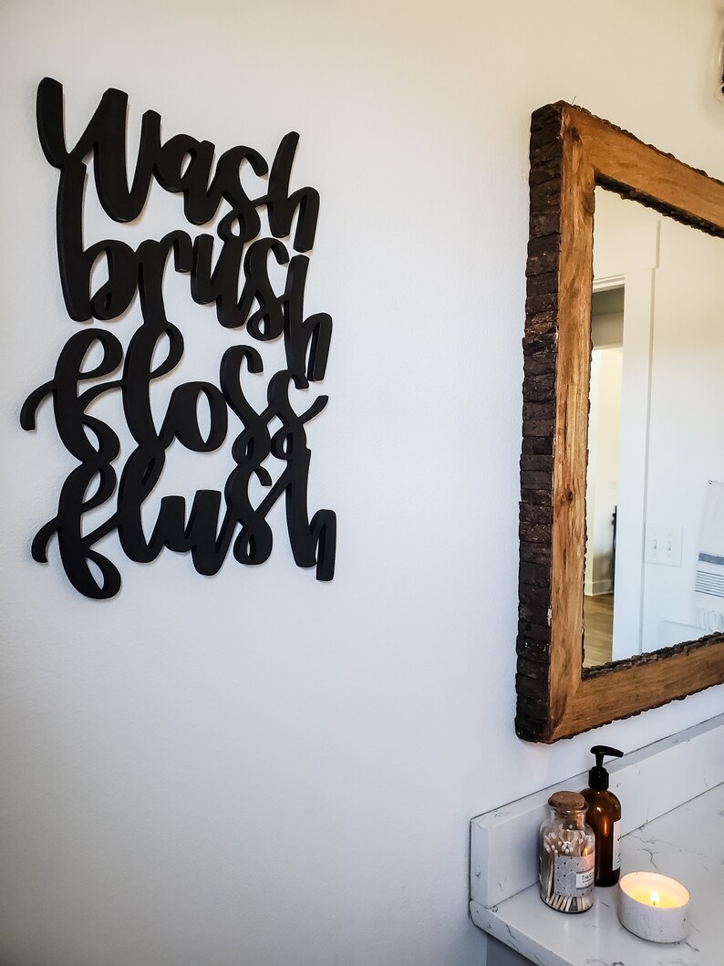 May include: Black wooden sign with the words "Wash, Brush, Floss, Flush" in a stylized font. The sign is hanging on a white wall next to a wooden framed mirror.