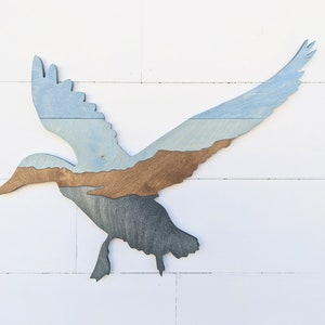 May include: A wooden silhouette of a duck in flight, painted in shades of blue and brown. The duck's wings are spread wide, and its body is angled downwards. The wood grain is visible in the paint.