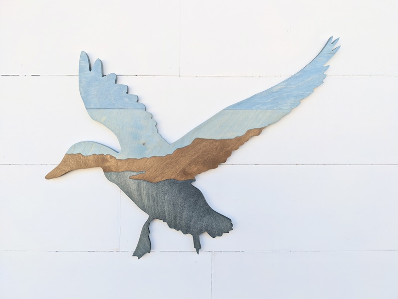 May include: A wooden silhouette of a duck in flight, painted in shades of blue and brown. The duck's wings are spread wide, and its body is angled downwards. The wood grain is visible in the paint.