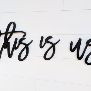 May include: Black wooden wall art spelling out "this is us" in a cursive font. The letters are separate and mounted on a white, horizontal-planked wall. The artwork is a decorative home accent.