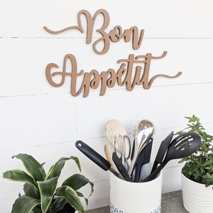 May include: A wooden sign with the words "Bon Appetit" in cursive script, hanging on a white wall. A white ceramic utensil holder with blue and white designs holds a variety of kitchen utensils, including wooden spoons, spatulas, and a whisk. Two potted plants with green leaves are on either side of the utensil holder.