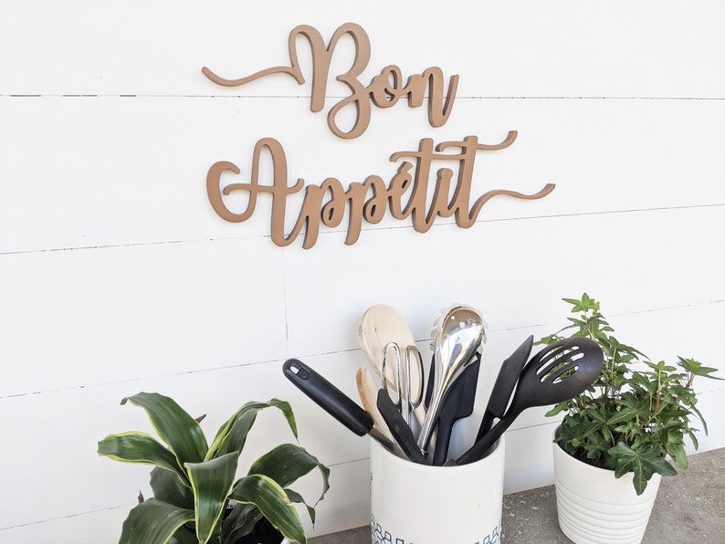 May include: A wooden sign with the words "Bon Appetit" in cursive script, hanging on a white wall. A white ceramic utensil holder with blue and white designs holds a variety of kitchen utensils, including wooden spoons, spatulas, and a whisk. Two potted plants with green leaves are on either side of the utensil holder.