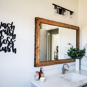 May include: A bathroom vanity with a white countertop and a white porcelain sink. A wooden framed mirror with a rustic wood frame hangs above the sink. A black sign with the words "Wash brush floss flush" is on the wall to the left of the mirror.