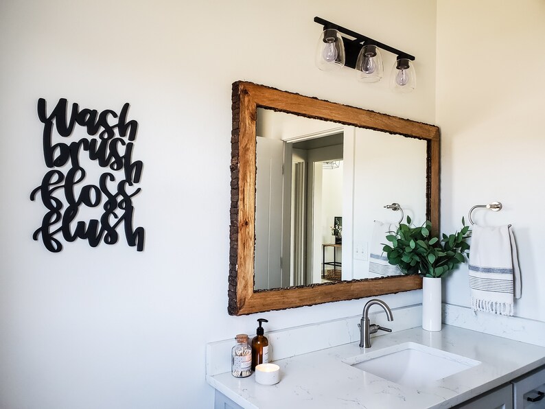 May include: A bathroom vanity with a white countertop and a white porcelain sink. A wooden framed mirror with a rustic wood frame hangs above the sink. A black sign with the words "Wash brush floss flush" is on the wall to the left of the mirror.