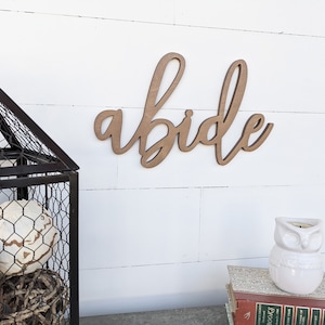 May include: Wooden sign with the word "abide" written in cursive script. The sign is brown and is mounted on a white wall. There is a black wire cage with white decorative balls in the lower left corner of the image. There is a white owl candle holder on a stack of books in the lower right corner of the image.