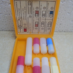 May include: A bright yellow plastic medical kit case containing various small cylindrical containers with colored caps. The interior lid features a printed instructional chart with Russian text and numbered diagrams for identifying the contents.