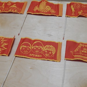 Peut inclure: Six drapeaux en tissu rouge avec des motifs jaunes. Les drapeaux présentent des images d'un soleil, d'une colombe, d'un navire, d'enfants, d'un bâtiment et d'une fille avec le texte "Пусть всегда будет солнце!" qui se traduit par "Que le soleil brille toujours !"