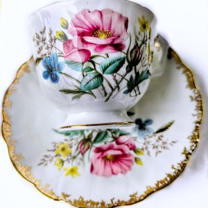 Op de afbeelding: Witte theekop en schotel set met een bloemenontwerp met roze rozen, blauwe bloemen en groene bladeren. De randen van de schotel en de basis van de kop zijn goudkleurig afgewerkt. Een klassieke theeset.