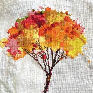 May include: Cross-stitch artwork of a tree with a brown trunk and branches. The tree's crown is a vibrant mix of orange, yellow, red, and pink, resembling autumn foliage. The artwork is on a white fabric background.