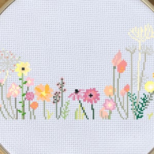 May include: Cross-stitch embroidery of a colorful wildflower meadow. The design features various flowers in shades of pink, yellow, orange, and green, set against a white background. The embroidery is framed in a round wooden hoop.