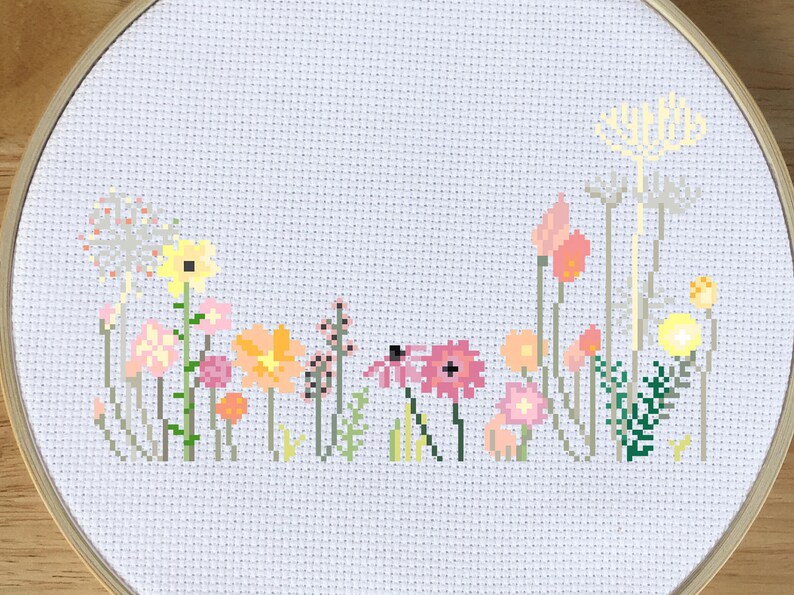 May include: Cross-stitch embroidery of a colorful wildflower meadow. The design features various flowers in shades of pink, yellow, orange, and green, set against a white background. The embroidery is framed in a round wooden hoop.