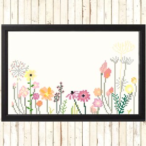 May include: A framed cross-stitch floral design. The artwork features a variety of pixelated flowers in shades of pink, yellow, orange, and green against a cream background. The frame is black, and the piece is displayed against a white wood-paneled wall.