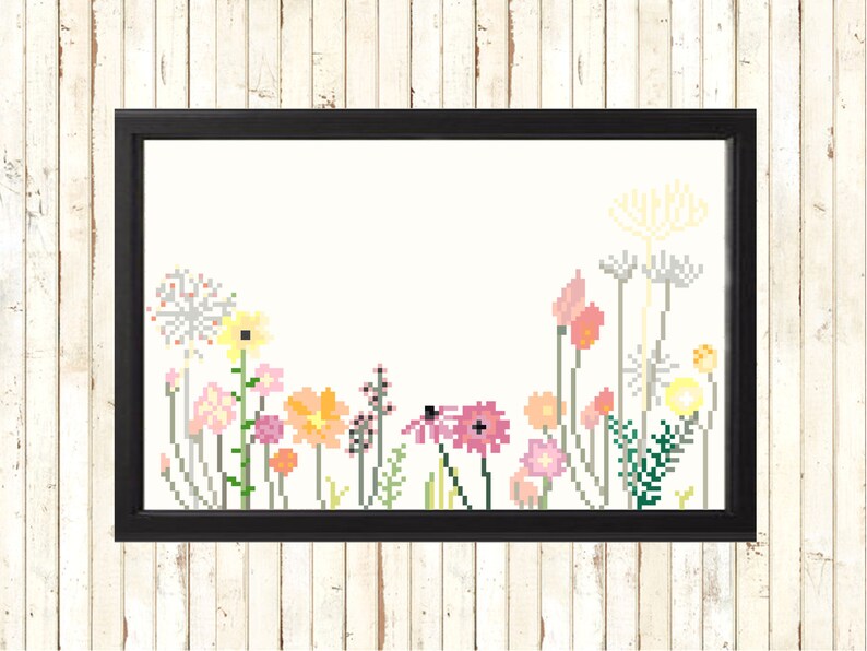 May include: A framed cross-stitch floral design. The artwork features a variety of pixelated flowers in shades of pink, yellow, orange, and green against a cream background. The frame is black, and the piece is displayed against a white wood-paneled wall.