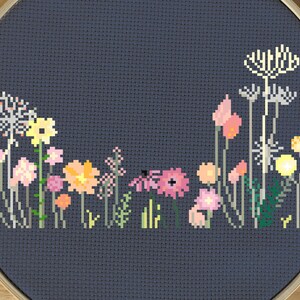 May include: A cross-stitch embroidery of a wildflower garden in a wooden hoop. The design features various flowers in shades of pink, yellow, and orange against a dark blue background. The embroidery is framed in a round wooden hoop.