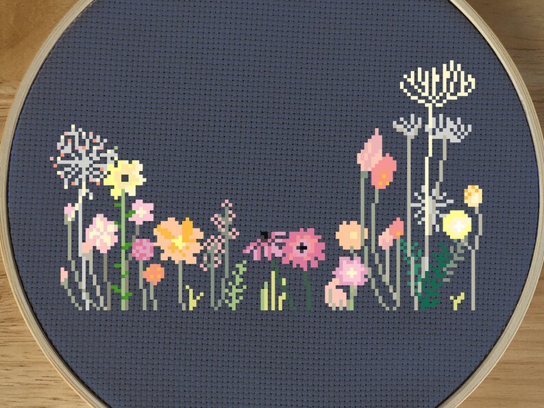 May include: A cross-stitch embroidery of a wildflower garden in a wooden hoop. The design features various flowers in shades of pink, yellow, and orange against a dark blue background. The embroidery is framed in a round wooden hoop.