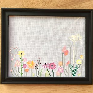 May include: A framed cross-stitch featuring a colorful floral design. The embroidery showcases various flowers in shades of pink, yellow, orange, and green against a white background. The frame is black, and the piece is displayed on a wooden surface.