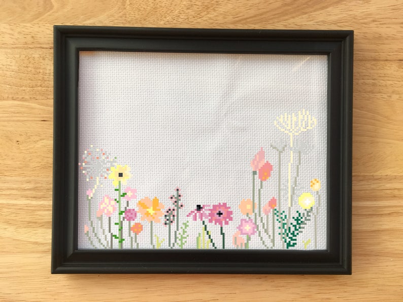 May include: A framed cross-stitch featuring a colorful floral design. The embroidery showcases various flowers in shades of pink, yellow, orange, and green against a white background. The frame is black, and the piece is displayed on a wooden surface.