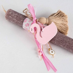 Può includere: Una candela grigia e bianca con una decorazione a forma di cigno rosa, un nastro rosa e bianco e un ciondolo a stella dorata.