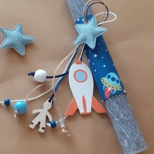 Può includere: Una candela pasquale grigia e bianca decorata con una stella di stoffa blu e bianca, un razzo, un disco volante e una ghirlanda di perline blu e bianche.