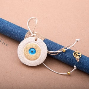 Può includere: Un rotolo di legno blu e bianco con un ciondolo in ceramica bianca con un occhio turco blu e dettagli dorati. Il ciondolo è attaccato al rotolo con uno spago e una corda bianca con perline dorate. Il rotolo ha il testo "my tree" scritto su di esso.