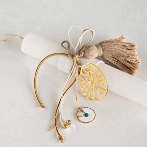 Può includere: Candela bianca con un ciondolo a forma di uovo in filigrana dorata, un ciondolo di occhio turco blu e un fiocco marrone. I ciondoli sono attaccati alla candela con nastri bianchi e marroni.